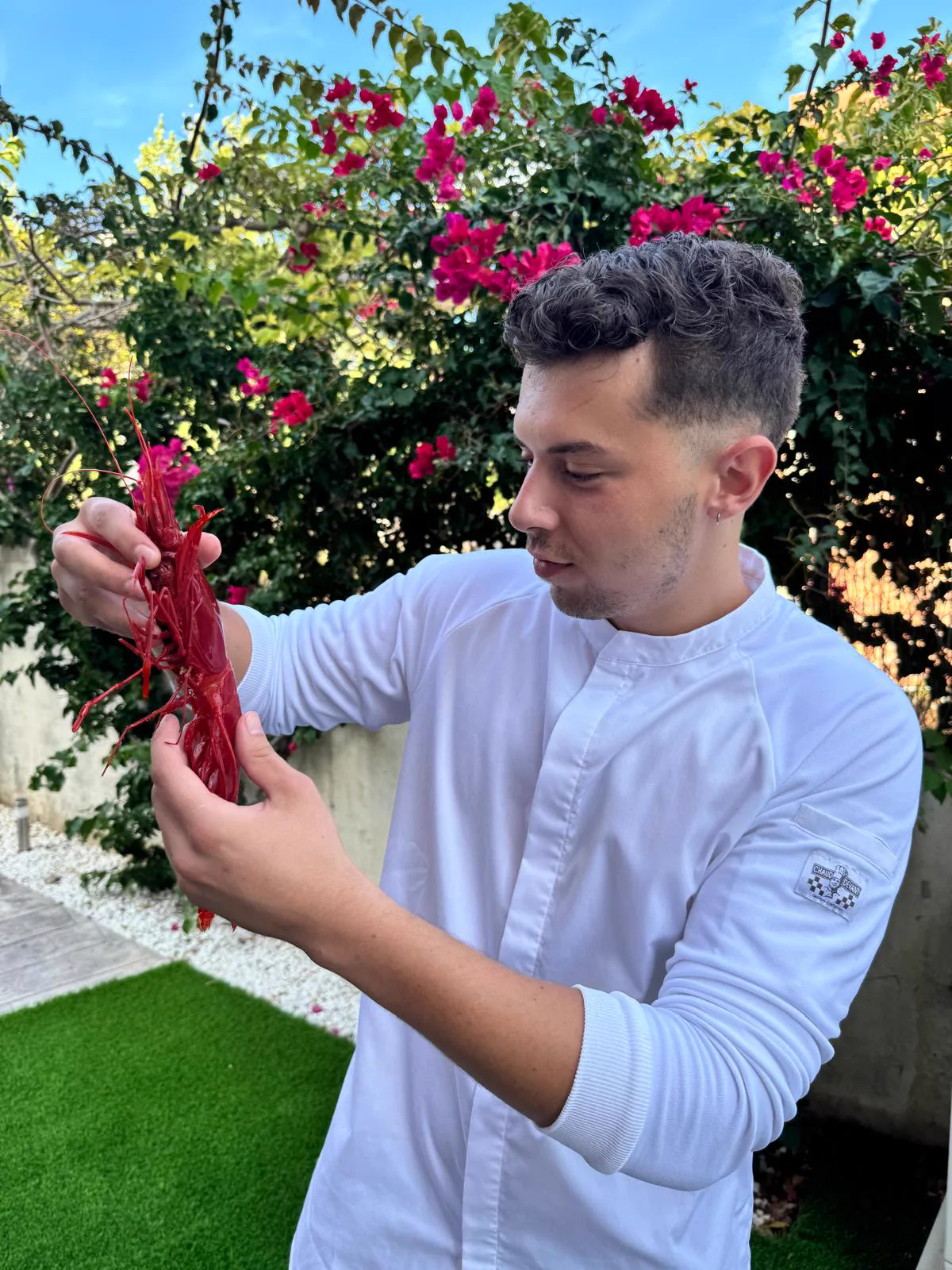 Sergi Molías in a white chef's jacket, holding a carabinero prawn in a garden with bougainvillea